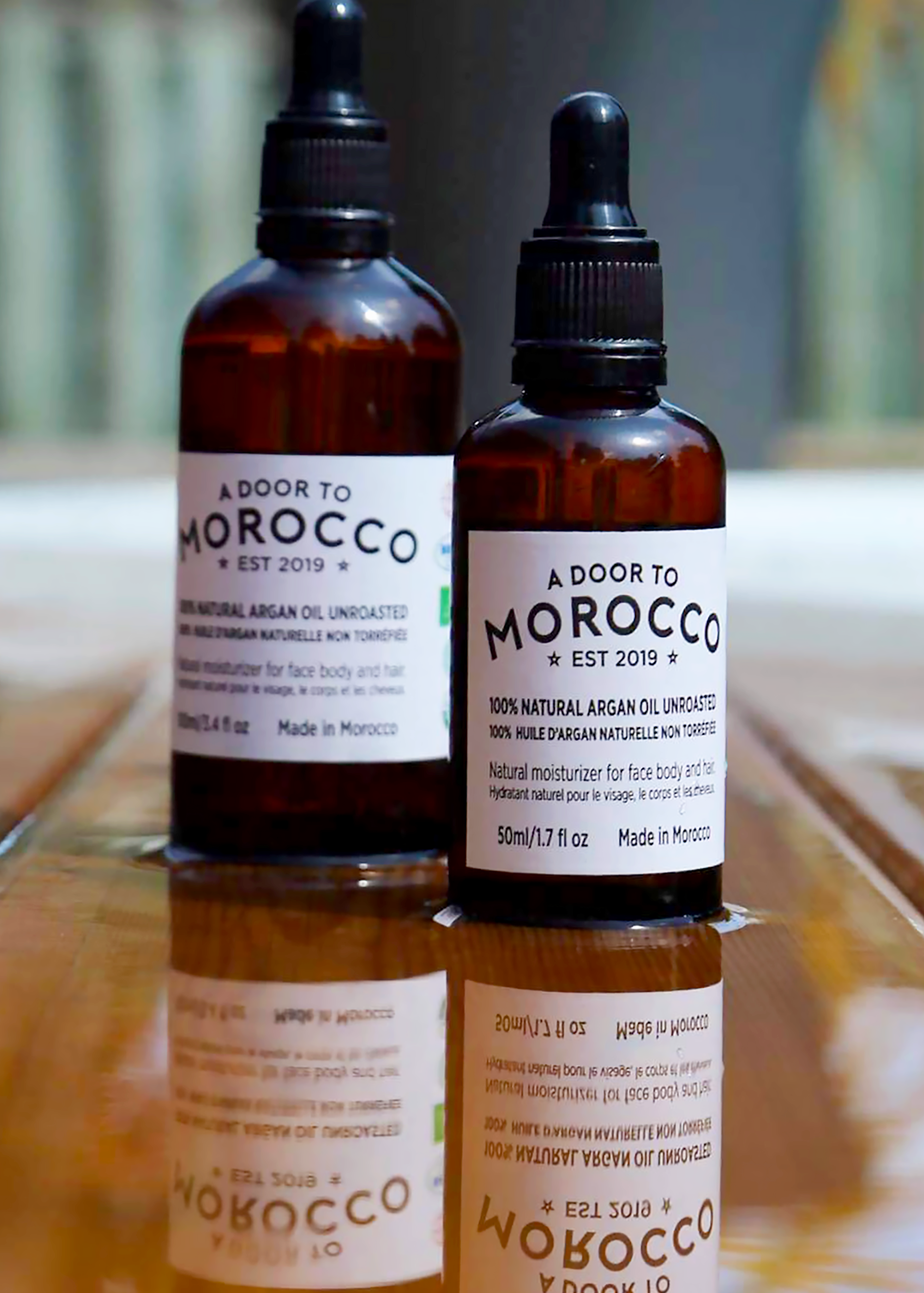 A Door to Morocco’s 50ml and 100ml bottles of argan oil with label sitting on wood with water displaying the reflection of bottles